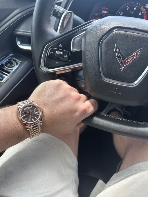 A close-up photo of a person's hand on the steering wheel of a Chevrolet Corvette (C7), wearing a rose gold Rolex watch with a black dial, with shorts visible and the car's tachometer in the background.