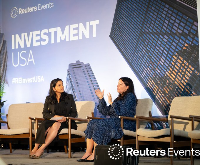 Two women sit on stage at Reuters Events' Investment USA conference, with one gesturing animatedly as she speaks to the other in front of a backdrop displaying "INVESTMENT USA" and "#REInvestUSA" against a cityscape image. The scene corresponds to the post, which notes that Grayscale's Head of Global Distribution, Laurie Katz, addressed an audience representing over $5 trillion in AUM at the event about institutional digital assets.