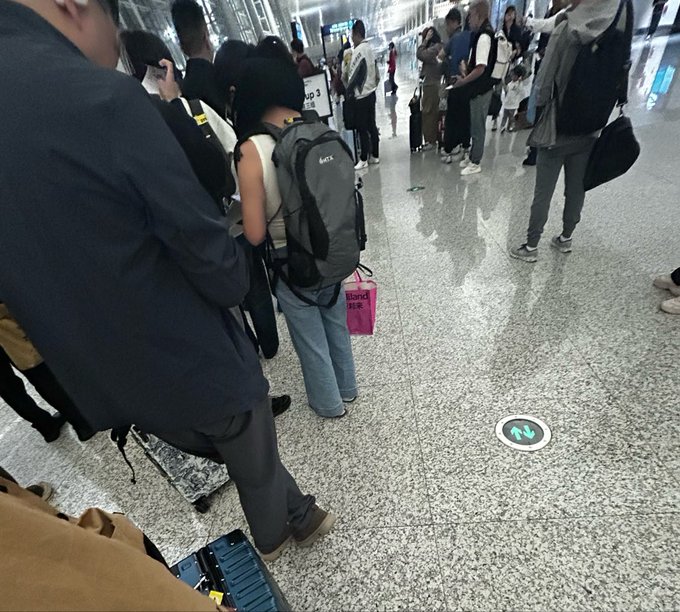 A traveler wearing a sleek gray backpack stands in an airport terminal, with other passengers and luggage visible around the polished floor featuring a directional arrow decal.

The image relates to the post by showcasing the stylish gray backpack that caught the traveler's eye, highlighting its appeal as a desirable travel accessory.