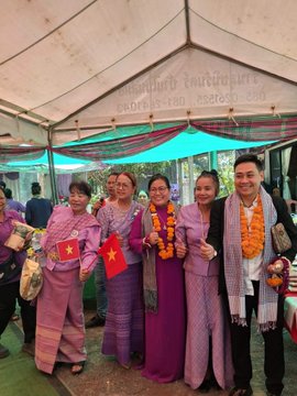 A group of five people, four women in traditional purple Lao/Thai dresses and one man in a suit, pose together under a tent wearing yellow flower garlands and holding small red flags with yellow stars.