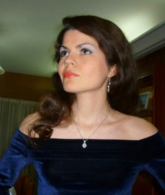 A woman with long brown hair styled in a classic 1960s look, wearing a dark blue velvet off-the-shoulder dress and pearl drop earrings, looks over her shoulder with a serious expression.