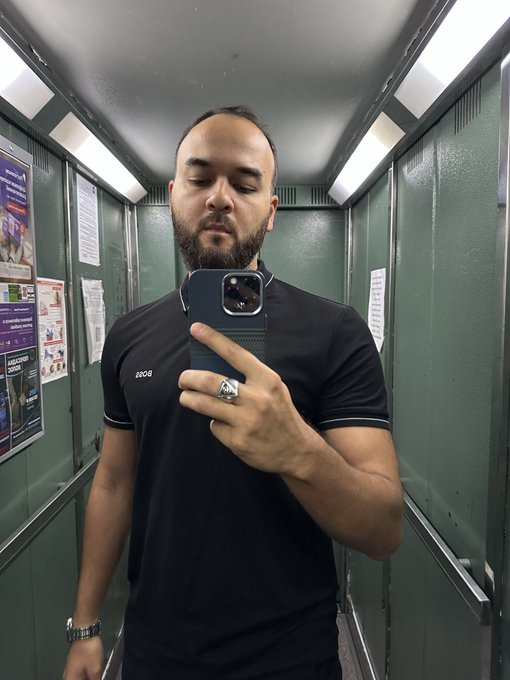 A man taking a selfie of himself inside a subway train, wearing a black shirt with his arm outstretched in front of him while holding up an iPhone camera towards the mirror.