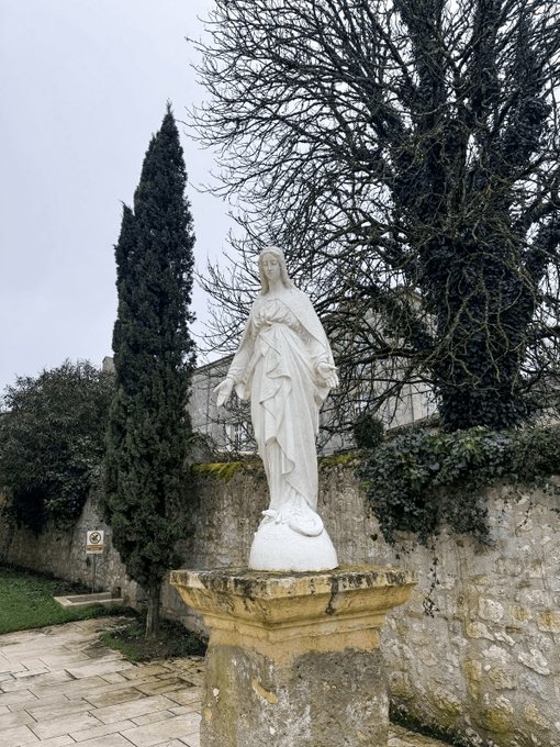 A white statue of the Virgin Mary stands on a stone pedestal beside a tall cypress tree against an old stone wall.