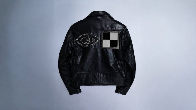 A product photograph of the back of a black leather jacket laid flat against a light gray background, decorated with silver studs forming an eye shape and a checkered square pattern.