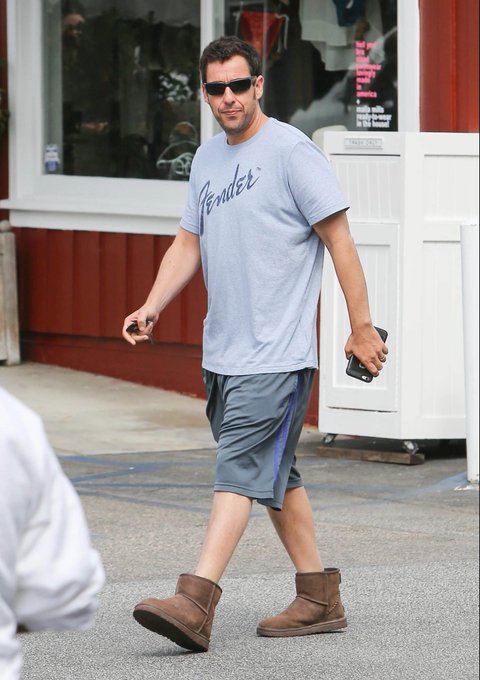 Adam Sandler walks outdoors wearing a gray Fender t-shirt, gray shorts, brown UGG boots, and sunglasses, while holding a phone.