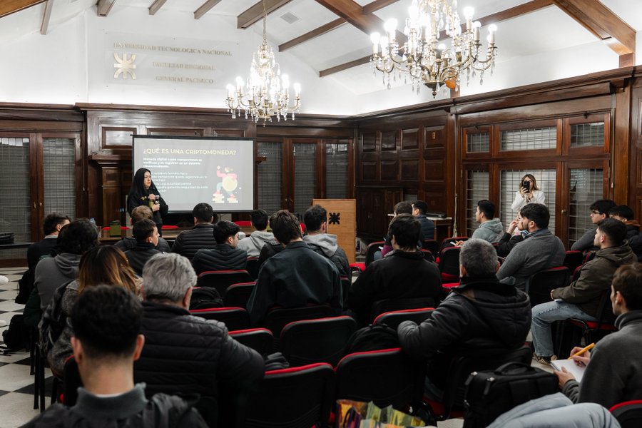 A presentation titled "¿Qué es una criptomoneda?" takes place in an elegant, wood-paneled hall at Universidad Tecnológica Nacional (Facultad Regional General Pacheco), where two female speakers address a seated audience of students during the Binance Academy University Tour stop in Buenos Aires, Argentina.
