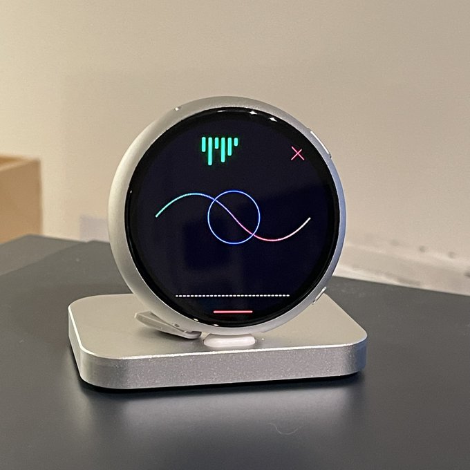 A round, silver smart display device with a dark circular screen showing a minimalist teal and pink abstract face design, sitting on a metal stand on a black surface.