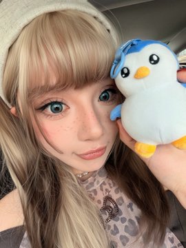 A young woman with long blonde hair, bangs, and a white knit hat poses for a selfie inside a car while holding up a blue and white Pudgy Penguins plush toy next to her face. The image accompanies a cheerful "Pengu Morning!!" post expressing excitement about bringing her Pudgy Penguins plush along for the day.