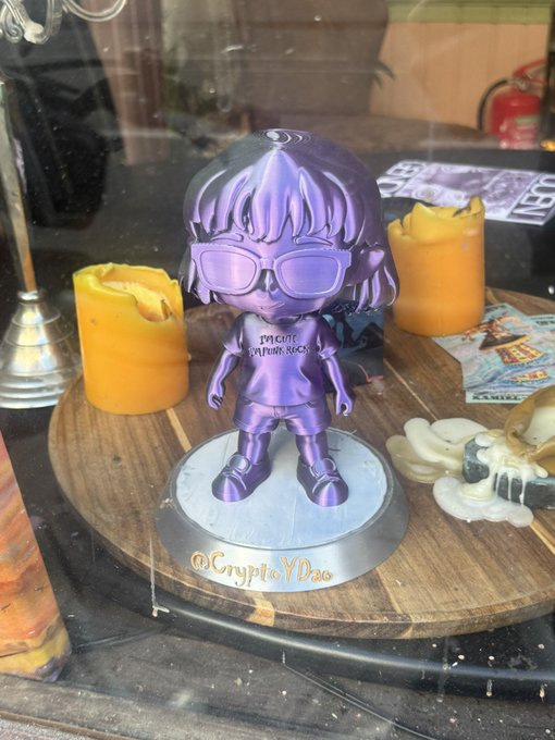 A metallic purple crypto-themed figurine of a person with sunglasses and a backpack, labeled 'Precise Capture Neck' and '@CryptoYDao' on its base, is displayed in a glass case near orange candles.