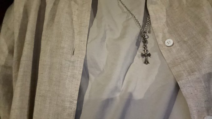 A close-up shot shows a silver cross pendant necklace worn over a white button-up shirt, partially covered by a light-colored cardigan.
