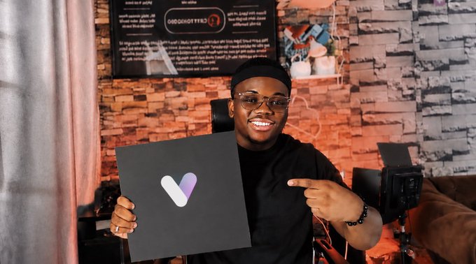 A smiling young man wearing glasses and a black shirt points to a black sign displaying a white and purple "V" logo, likely for VeWorld, inside a studio with a brick-patterned wall.