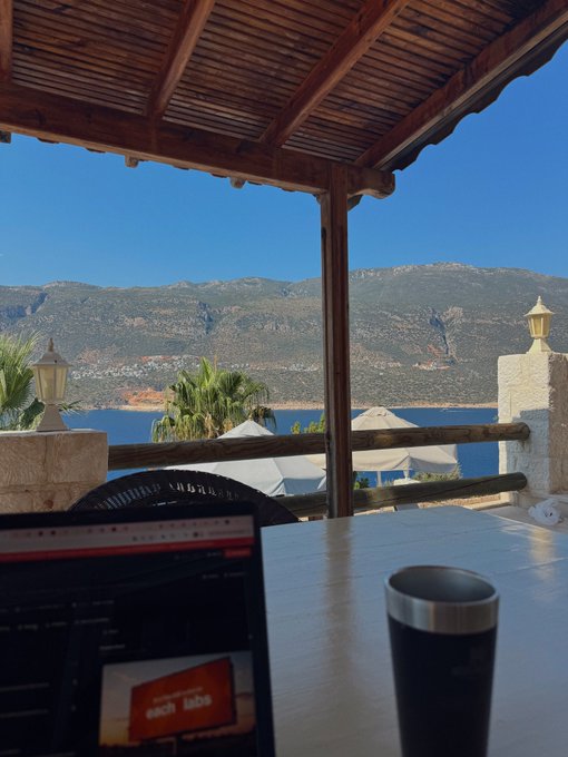 A scenic view from a wooden-roofed patio overlooking a blue sea and mountains, with a laptop and a black travel tumbler resting on a white table in the foreground.