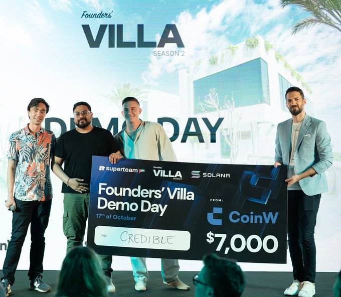 Four people stand on stage at the Founders' Villa Season 2 Demo Day, holding a large $7,000 check from CoinW presented to "Credible" alongside Superteam, Villa, and Solana branding. The image captures the team's early 2024 milestone, which the post contrasts with their later $32M MetaDAO raise in 2026 to highlight their growth.
