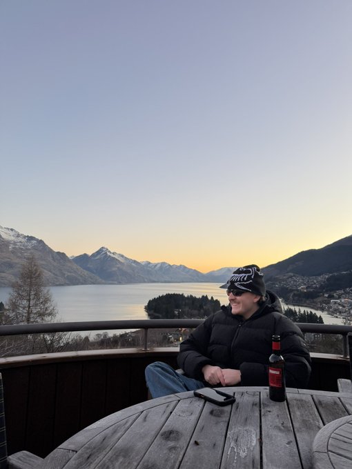 A man wearing a beanie and puffer jacket sits at a wooden table with a beer bottle, overlooking a serene mountain lake at sunset. The scenic alpine backdrop with snow-capped peaks complements the casual vibe, making it the perfect setting to ask "who wants $ANSEM."