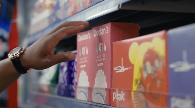 A hand wearing a gold watch reaches toward boxes of 'Declan's White Rice' on a grocery store shelf alongside other 'Peak' brand products.