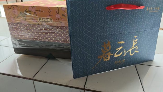 Two decorative boxes sit on a tiled floor; the right one is a dark blue gift bag featuring gold Chinese calligraphy reading '譽云長' and a red ribbon handle, and the left is a wrapped gift box with floral designs.