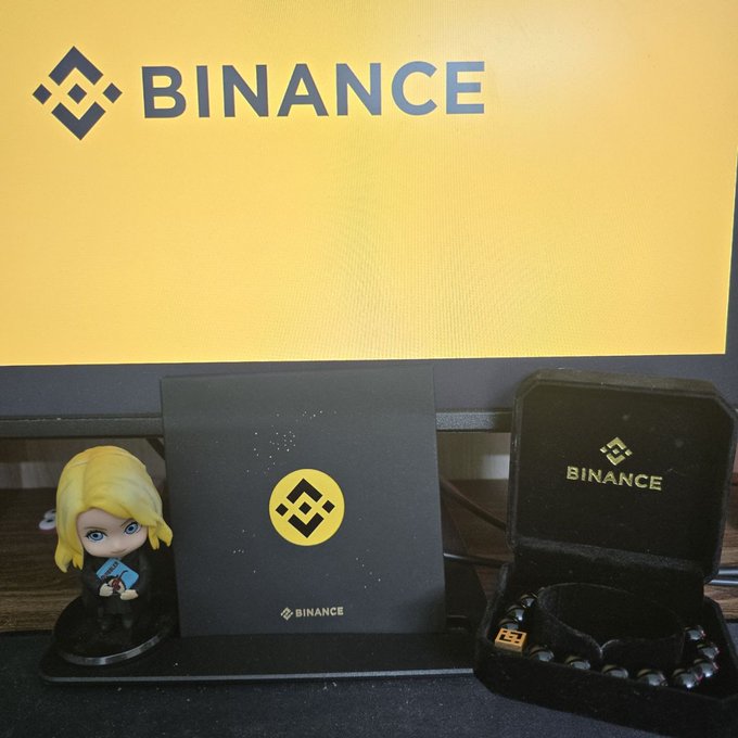 A Binance-themed display featuring the company's logo on a yellow screen, accompanied by a blonde figurine, a black Binance-branded card with a gold coin emblem, and an open jewelry box containing a black beaded bracelet with the Binance logo charm — showcasing the thoughtful gift mentioned in the post.