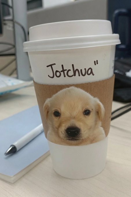 A white coffee cup with a brown sleeve featuring an adorable golden retriever puppy, labeled "Jotchvä" in handwritten text, sits on a desk beside a pen and notebook. The post's "Wake up and smell the coffee" message pairs perfectly with the cozy caffeine companion, while "ILY" (I love you) is echoed by the irresistibly cute puppy peeking out from the cup.