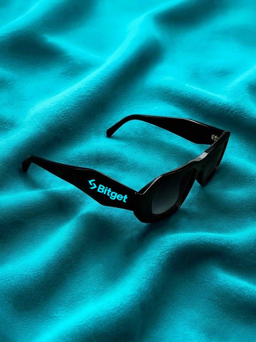 Black sunglasses branded with the 'Bitget' logo rest on draped turquoise satin fabric.