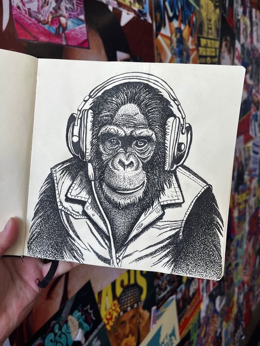 A hand holds open a sketchbook displaying a detailed pen-and-ink drawing of a gorilla wearing headphones and a jacket, set against a colorful poster-covered wall.