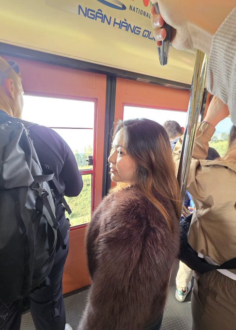 Inside a cable car or funicular with Vietnamese text 'NGÂN HÀNG ĐÔNG' visible on a sign, featuring a woman with long brown hair wearing a fuzzy coat looking out the window.