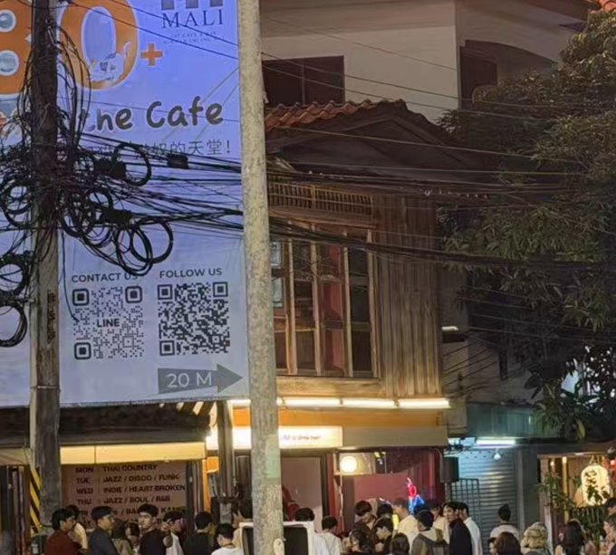 A nighttime street scene shows a crowd gathered outside a wooden building with a sign reading 'The Cafe' and Chinese characters, featuring QR codes for LINE contact and following, with a '20 M' directional arrow below.