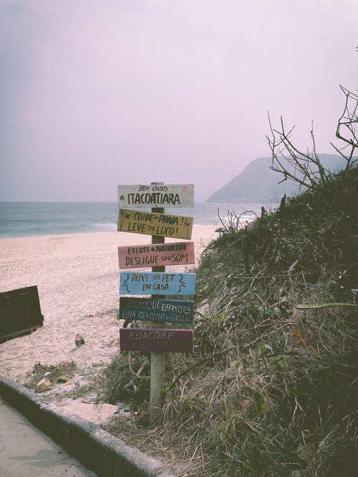 A weathered wooden signpost on a sandy beach displays stacked painted signs in Portuguese, including text reading "BEM VINDOS A ITACOTIARA," "CUIDADO COM A LIXO!," and "DUQUE SUAVE EM CASA," with the ocean and a distant mountain in the background.