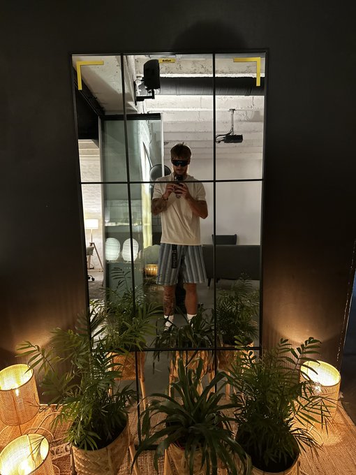 Mirror selfie of a man wearing sunglasses, a cream t-shirt, and plaid shorts, reflected in a tall mirror surrounded by potted plants and warm ambient lighting.