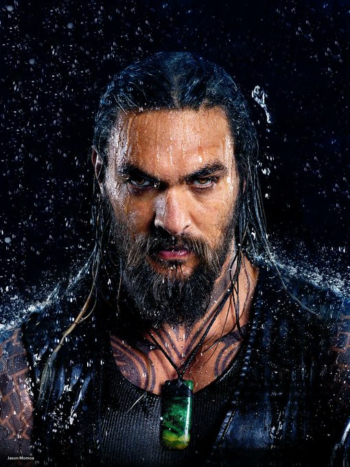 A dramatic portrait of actor Jason Momoa as Aquaman, with wet hair and beard, intense gaze, tribal tattoos, and a green pendant against a dark background with water splashing.