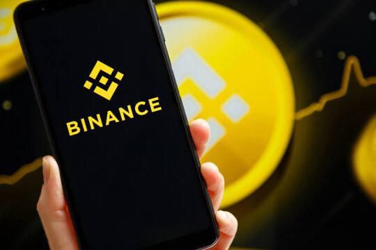 The image features a person holding up a smartphone displaying the binance logo. The background of the photo has lines that might be interpreted as cryptocurrency symbols, suggesting that the scene could have a connection with Bitcoin or other cryptocurrency-related services. The main focus of the image is the smartphone and its content, which appears to be related to the post's claim about Binance blurring the line between stocks and crypto.