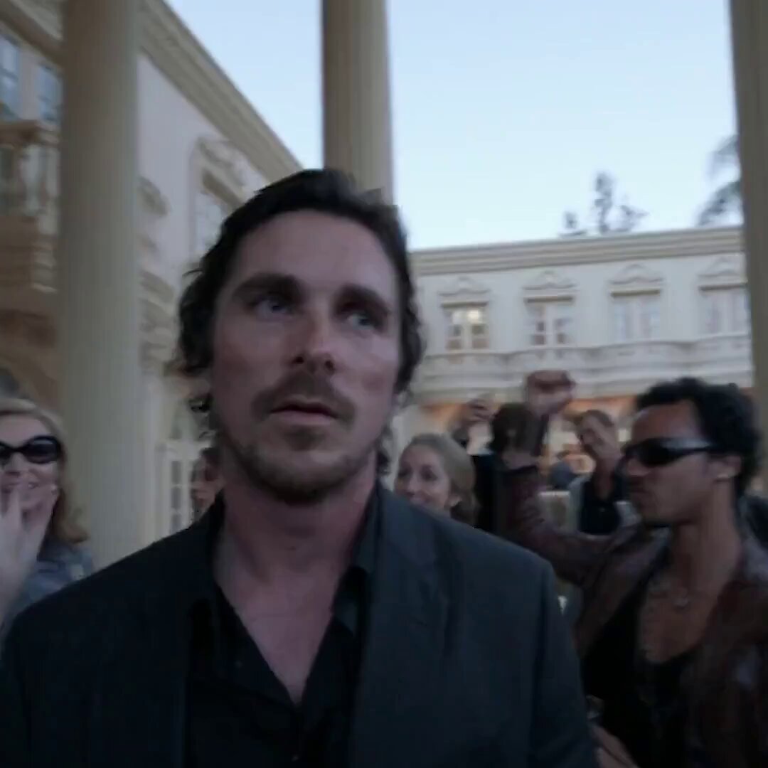 The image shows a man wearing a suit standing outside of an ornate building, surrounded by several people who are also dressed up. The group appears to be attending a formal event or gathering. One person in the crowd has their hand raised, possibly addressing someone or making a point during the event.