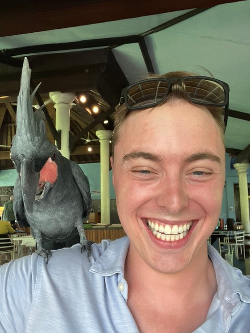 A smiling young man with sunglasses on his head takes a selfie while a palm cockatoo perches on his shoulder at what appears to be a restaurant.