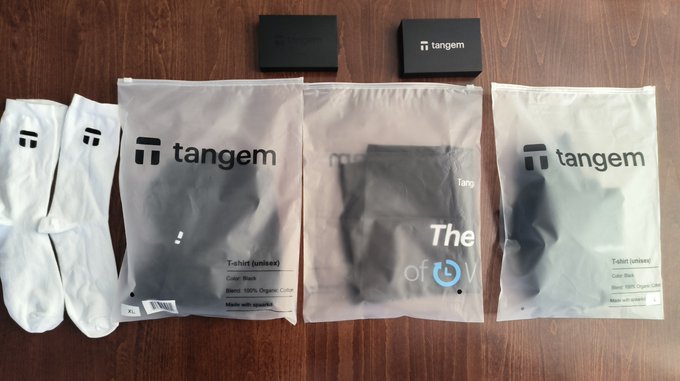 A pair of white socks and several sealed plastic packages from Tangem are laid out on a wooden surface, including items labeled "T-shirt (unisex)" in various colors. The image relates to the post by showcasing the merchandise received as gifts, with the packages containing apparel featuring the Tangem brand name.