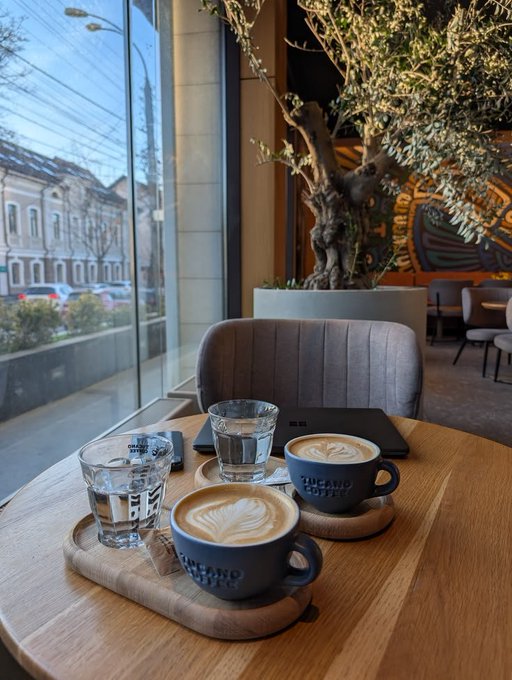 Two lattes with leaf latte art and glasses of water sit on a wooden table in a cozy café with an olive tree and window view of the street, with "THE AND COFFEE" branding visible on the cups — a perfect "good morning" coffee ritual to start the day.