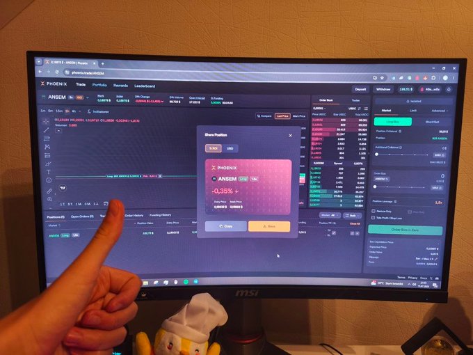 A person's hand giving a thumbs-up in front of an MSI monitor displaying the Phoenix trading platform with a share position dialog for ANSEM showing -0.35%.