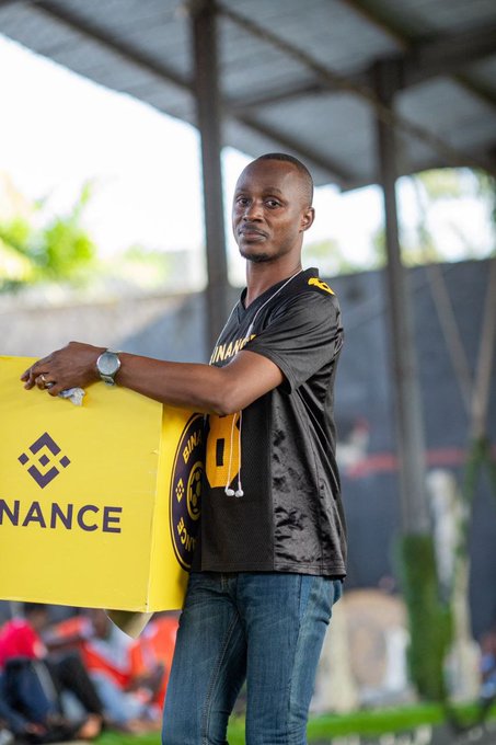 A man in a dark t-shirt and jeans stands under a shelter holding a yellow Binance promotional banner, with a wristwatch visible on his arm.