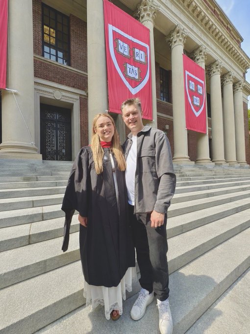 In this image, there are two people posing for a picture outside a building with red banners hanging from its columns. The person on the left side of the photo is wearing a black graduation gown, while the other person standing next to them has a gray jacket on. They both have smiles on their faces as they pose together in front of the Harvard building.