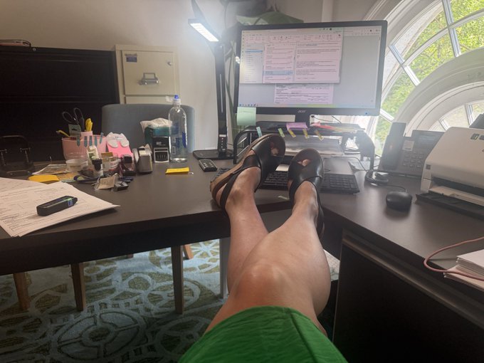 A first-person view shows a person's legs propped up on a cluttered office desk with a computer monitor, water bottle, and office supplies.