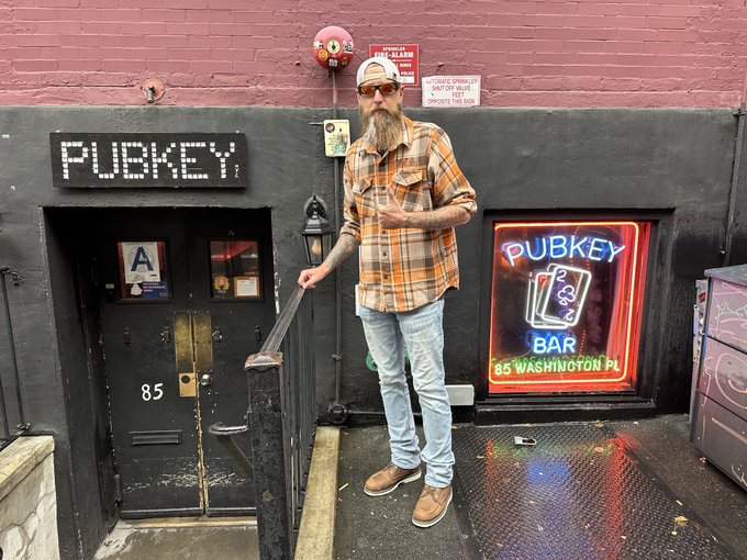 A bearded man wearing sunglasses, a plaid shirt, and a cap stands at the entrance of Pubkey, a bar located at 85 Washington Place, with a neon sign displaying the establishment's name and address visible behind him.