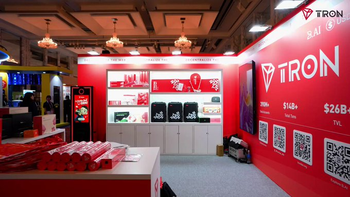 A TRON booth at a conference showcases the blockchain's brand with prominent red displays featuring the TRON logo, statistics like "200M+ Total Accounts," "$14B+ Total Txn," and "$26B+ TVL," alongside QR codes and merchandise. The setup aligns with the post's theme by visually representing TRON's role in enabling high-frequency, low-cost transactions for an emerging agentic economy.