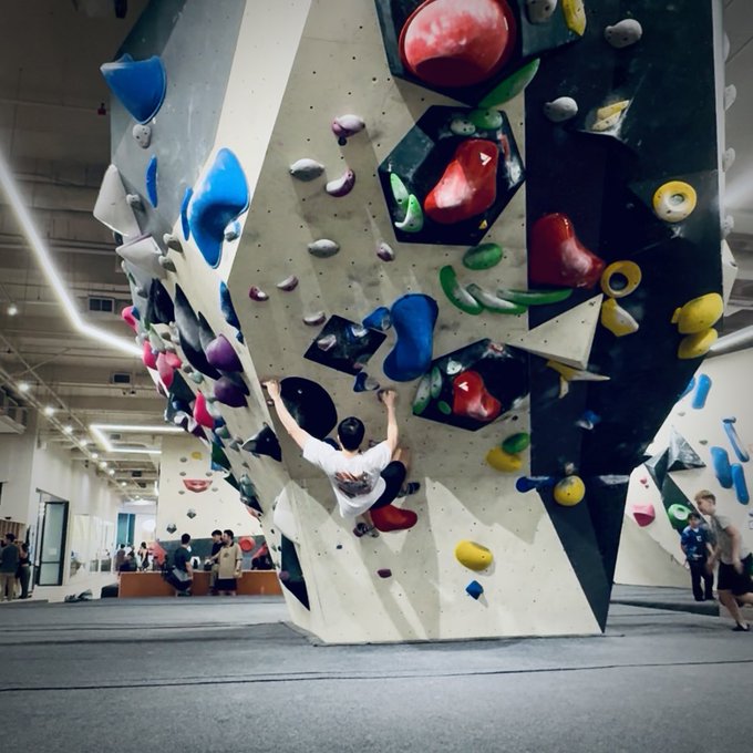 A young man in a white shirt scaling a colorful indoor bouldering wall dotted with multicolored holds.