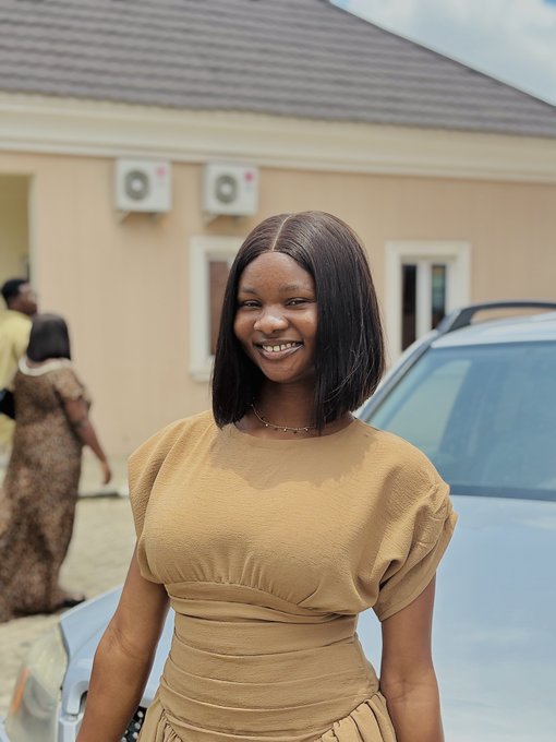 A young woman with shoulder-length dark hair smiles brightly while wearing a tan dress, standing outside a building with AC units mounted on the wall, likely preparing for an upcoming IRL event in Abuja as mentioned in her week to-do list.