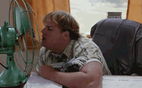 A heavyset man leans over a desk, blowing into the cage of a vintage mint-green electric fan as if singing into a microphone, while papers stamped with the name "ANGIE" lie in front of him. The staged moment visually echoes the caption's playful idea of "broadcasting" his presence to the world like a crypto announcement, with the fan standing in for a megaphone.