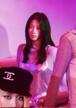 A photo with pink lighting showing a woman with long dark hair wearing a sparkly top, next to someone wearing a black Chanel hat.