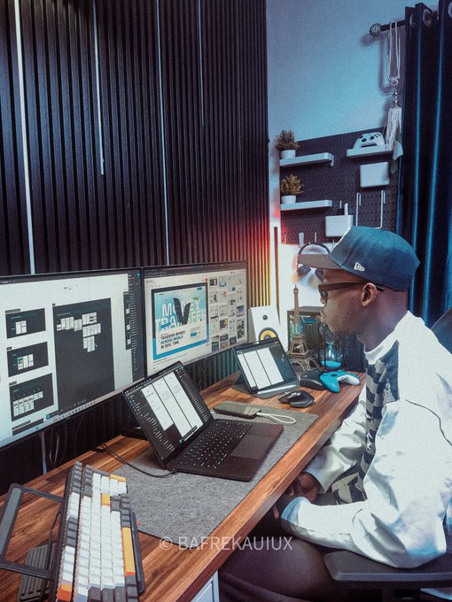 A photograph of a person in a white jacket and blue cap working at a multi-monitor desk setup with a laptop, mechanical keyboard, and a poster reading 'We Are Team-Based' on the wall, watermarked '@BAFREKAUIIUX'.