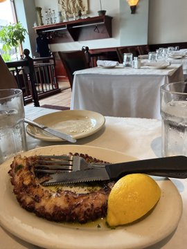 A close-up photo of a plated meal at a restaurant, featuring what appears to be a breaded cutlet or fish, accompanied by a lemon wedge, with utensils and a water glass visible on the white tablecloth.