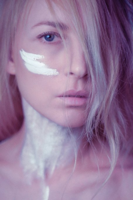 The image features a woman with blonde hair, wearing makeup that includes white eyeliner and lipstick. She has her hair styled in loose waves, giving her a casual yet stylish appearance. The background of the photo appears to be a light purple color, which complements the overall look of the woman's face. There is also an element of pinkish-purple tinting visible on the image, adding depth and dimension to the scene.