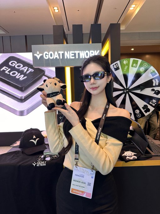 A young woman wearing sunglasses poses holding a GOAT NETWORK plush toy at a promotional booth, with a GOAT NETWORK sign, GOAT FLOW display, prize wheel, and merchandise visible. She wears a lanyard with a badge that appears to read 'REWARDS MEDIAWIKI JECIN' and 'token2049'.