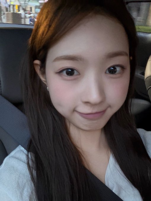 A selfie of a young East Asian woman with long brown hair wearing a white top, seated in a vehicle with a seatbelt visible.