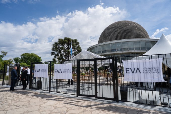 An outdoor event setup at a modern venue with a distinctive spherical mesh roof, featuring white banners reading 'EVA Ethereum Valuators Association' attached to black metal fencing.